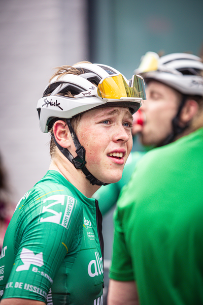 Two cyclists wearing helmets and green Wielrennen jerseys.