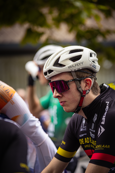 A man in a racing uniform with a white helmet is sitting on a bike with a black, blue and red jersey that says "De Ade".