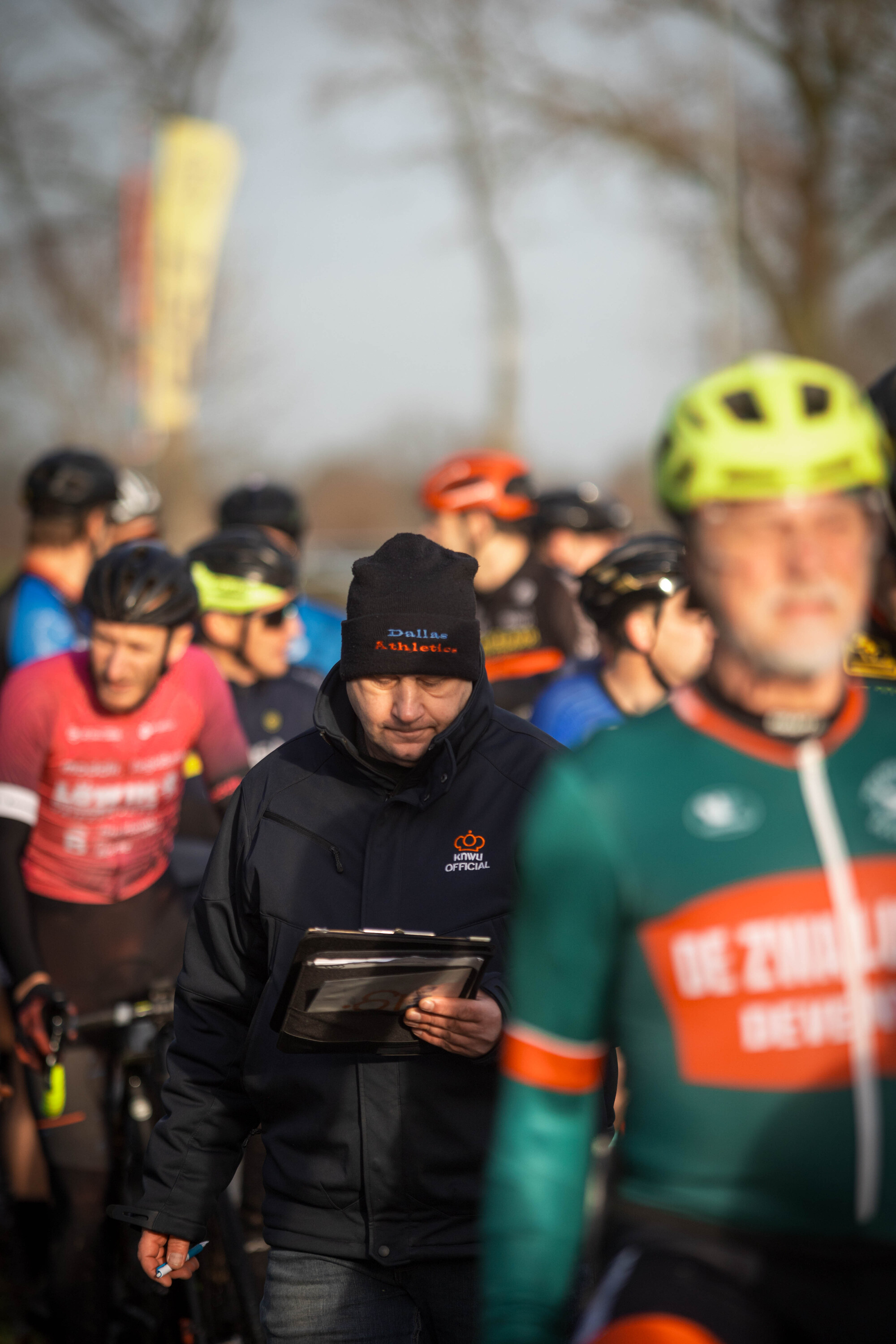 A man wears a green and red shirt with the word "Deventer" on it as he stands in front of other cyclists.