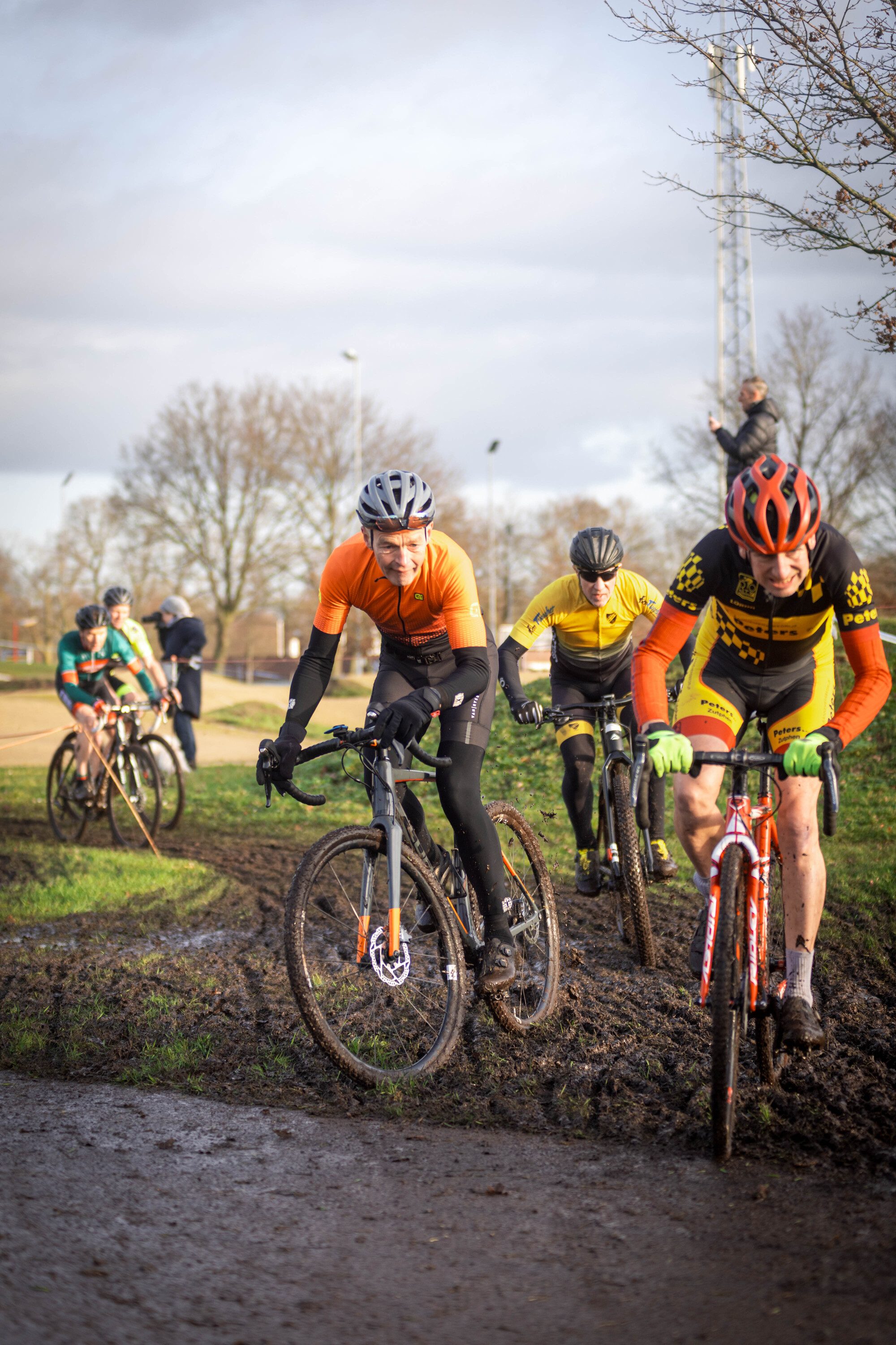 A man on a bike wearing a black helmet and an orange shirt is riding past another man who is also wearing a black helmet.