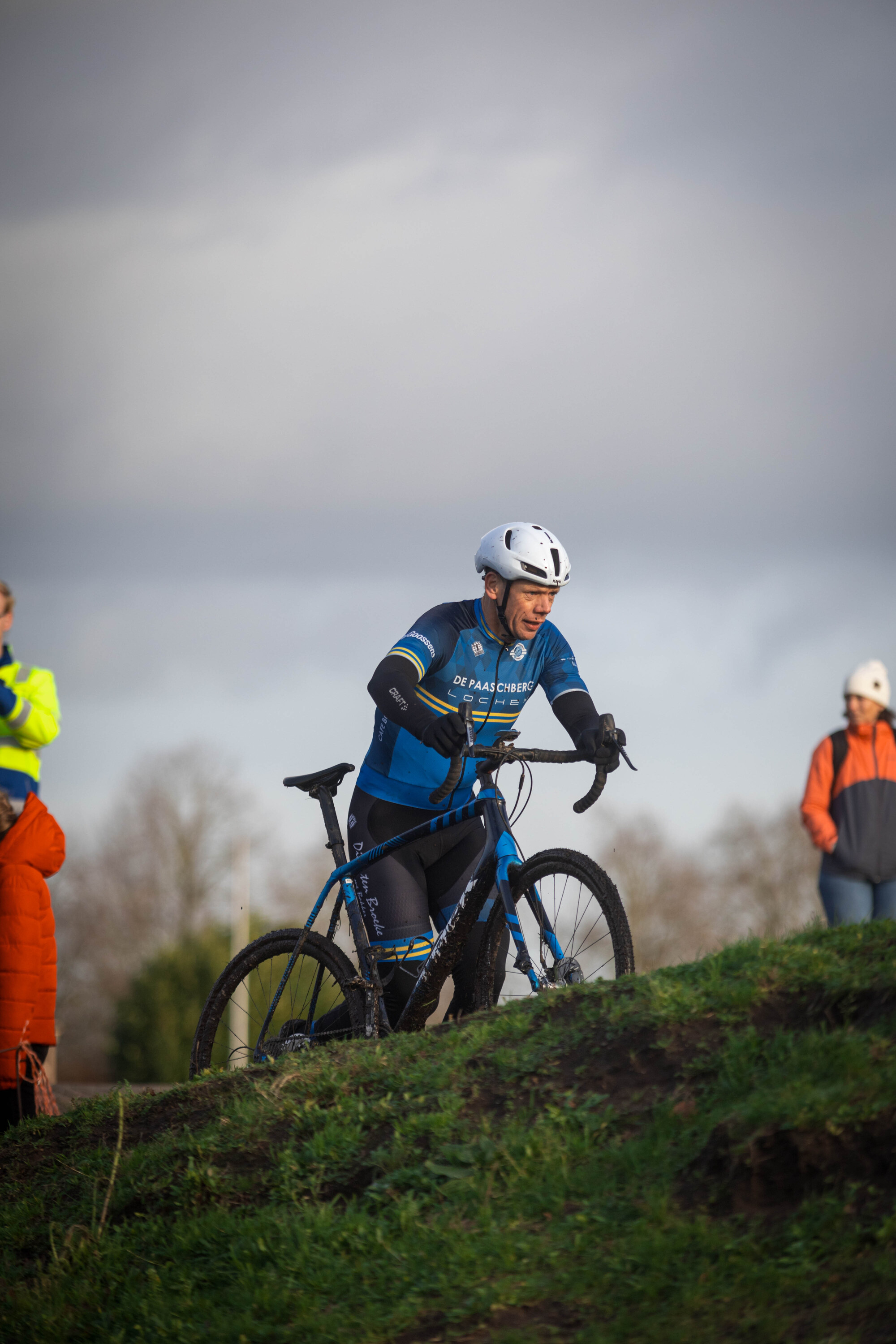 A man is riding a bike wearing an orange vest and black pants. He is standing next to another person on a mountain.