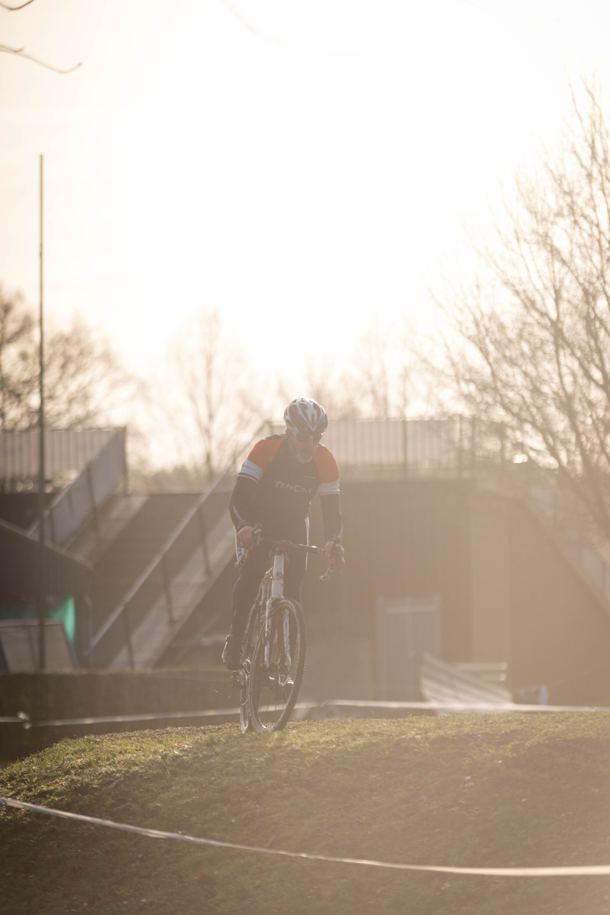 A man in a black and orange shirt is riding his bicycle on an uneven ground with a large building in the background.