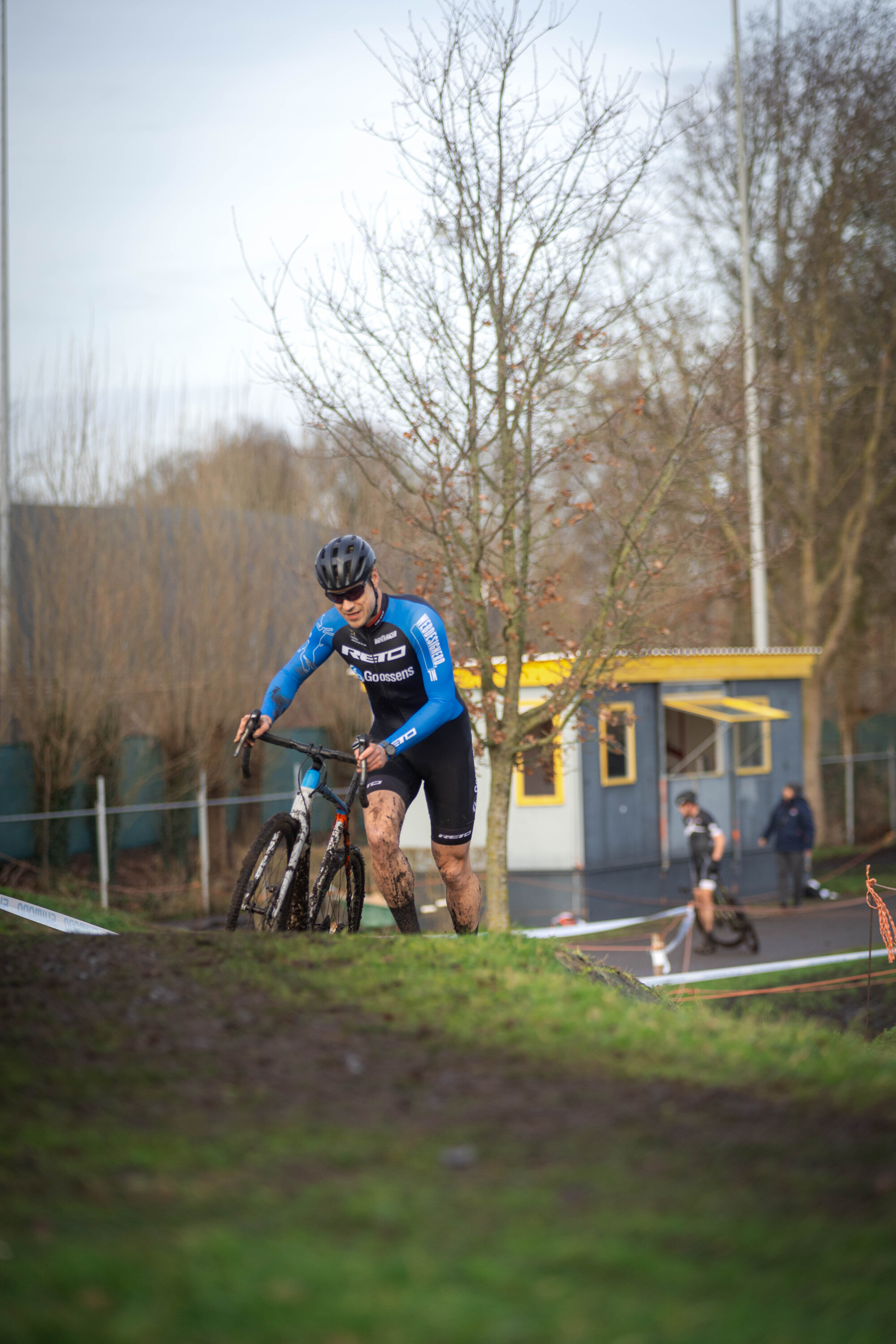 A cyclist wearing a blue and white suit is riding on a grassy hill in front of a small building.