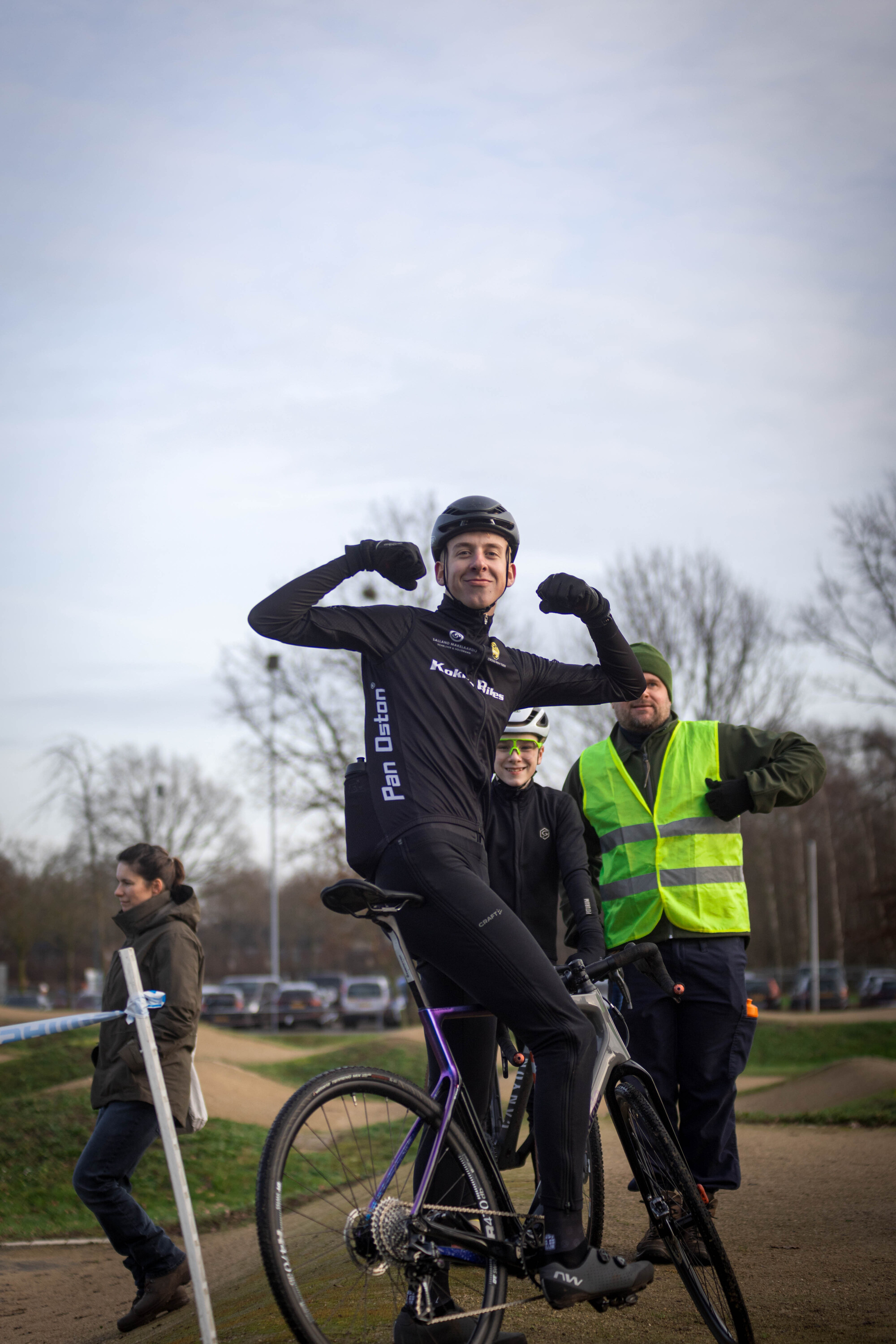 A man on a bike in black with the words "De Koert" written on his back.