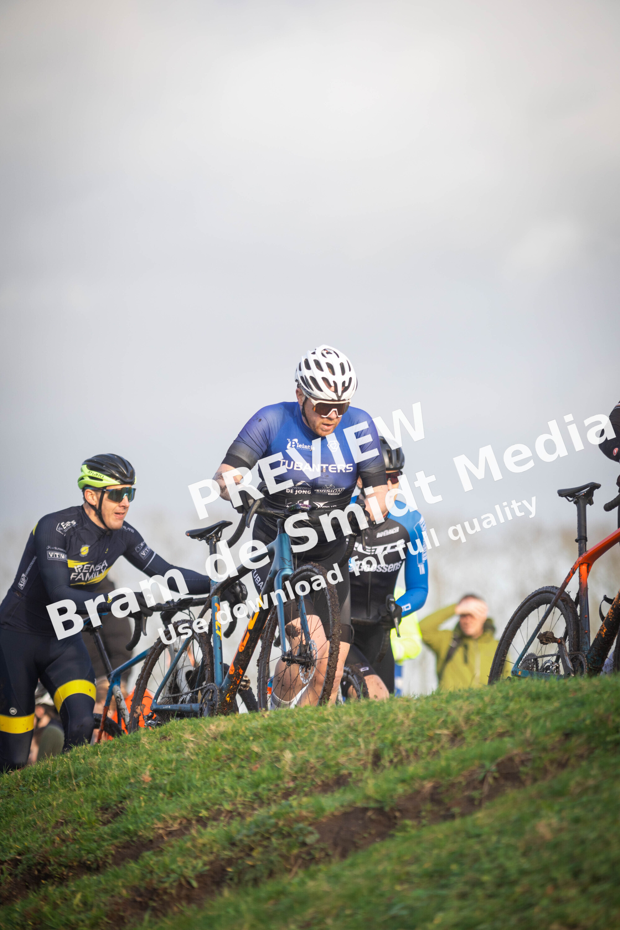 Three cyclists are seen on a grassy hill with one of them wearing a blue shirt and the number 2023.
