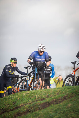 Three cyclists are seen on a grassy hill with one of them wearing a blue shirt and the number 2023.