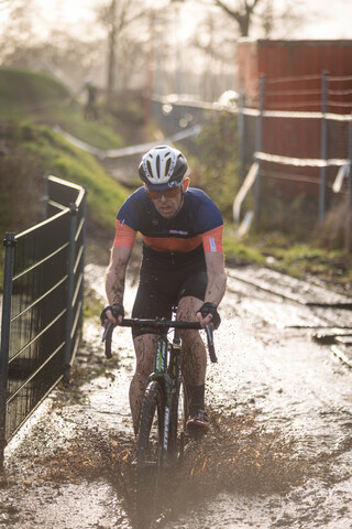 A cyclist in a blue and orange jersey is riding through mud. The rider's helmet features the letter M.