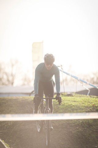 A cyclist in a green jacket and helmet riding on a bike with grass on the ground in the background.