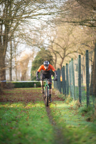 A man riding a green mountain bike on a path with trees and a wooden fence in the background. He is wearing an orange shirt.