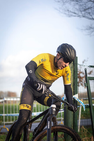 A man is riding a black bicycle in the rain while wearing a yellow shirt with GOW Raalte on it.