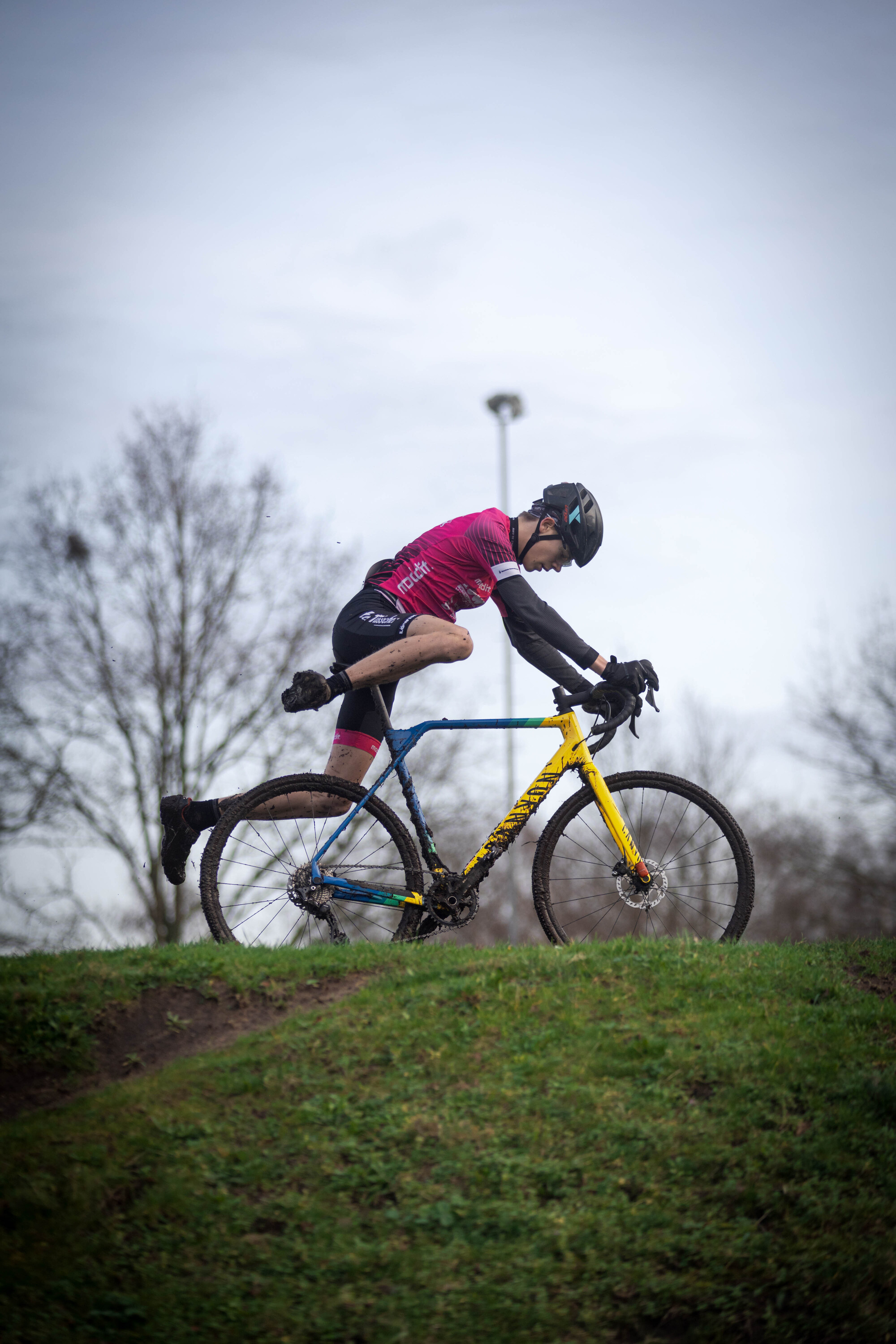 Cyclocross rider in a pink shirt and black gloves riding a blue bike with the word "JU/EL/BE/AM" on it.