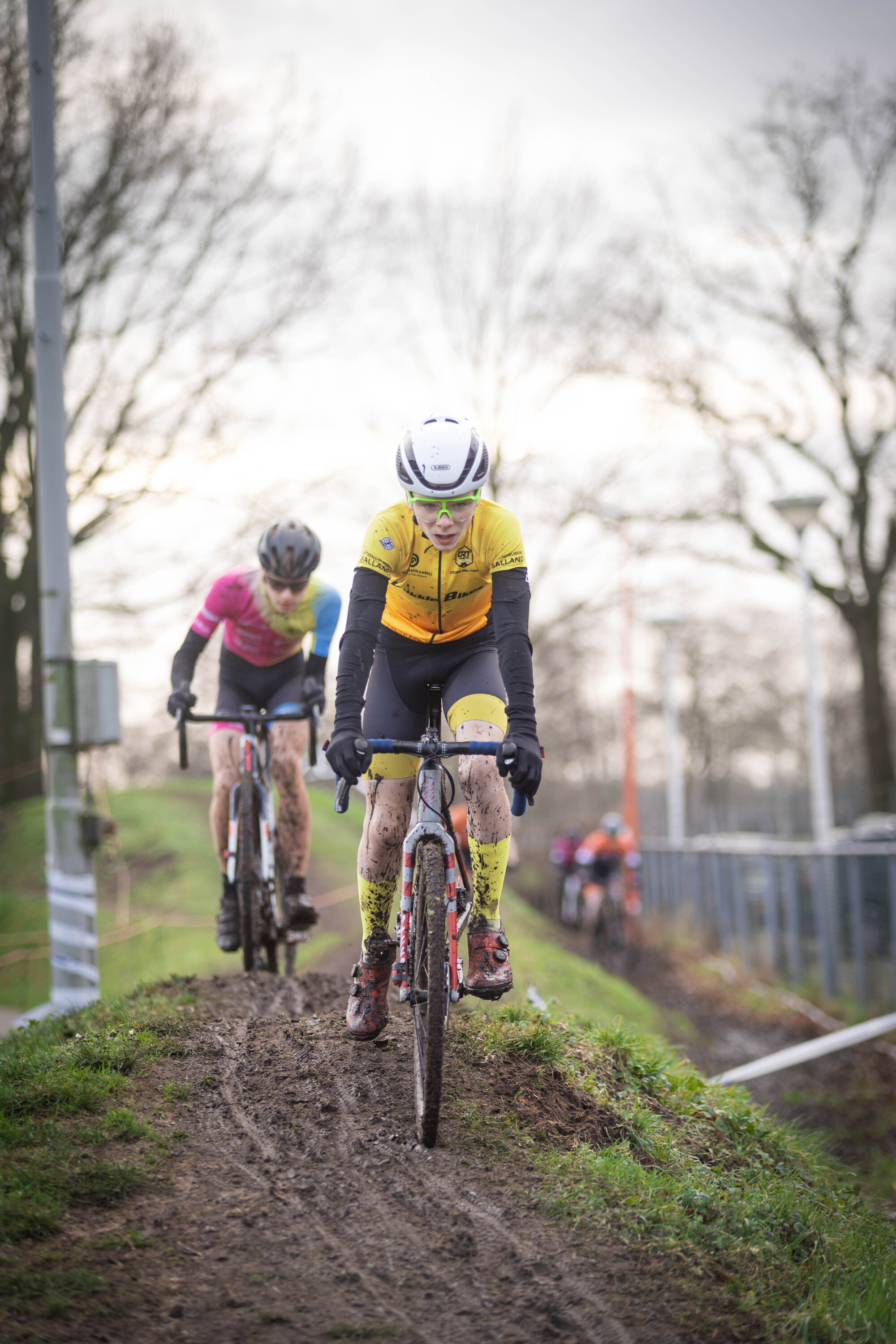 Two women are riding bikes up a hill. They have on uniforms that say Ju, BE and El.