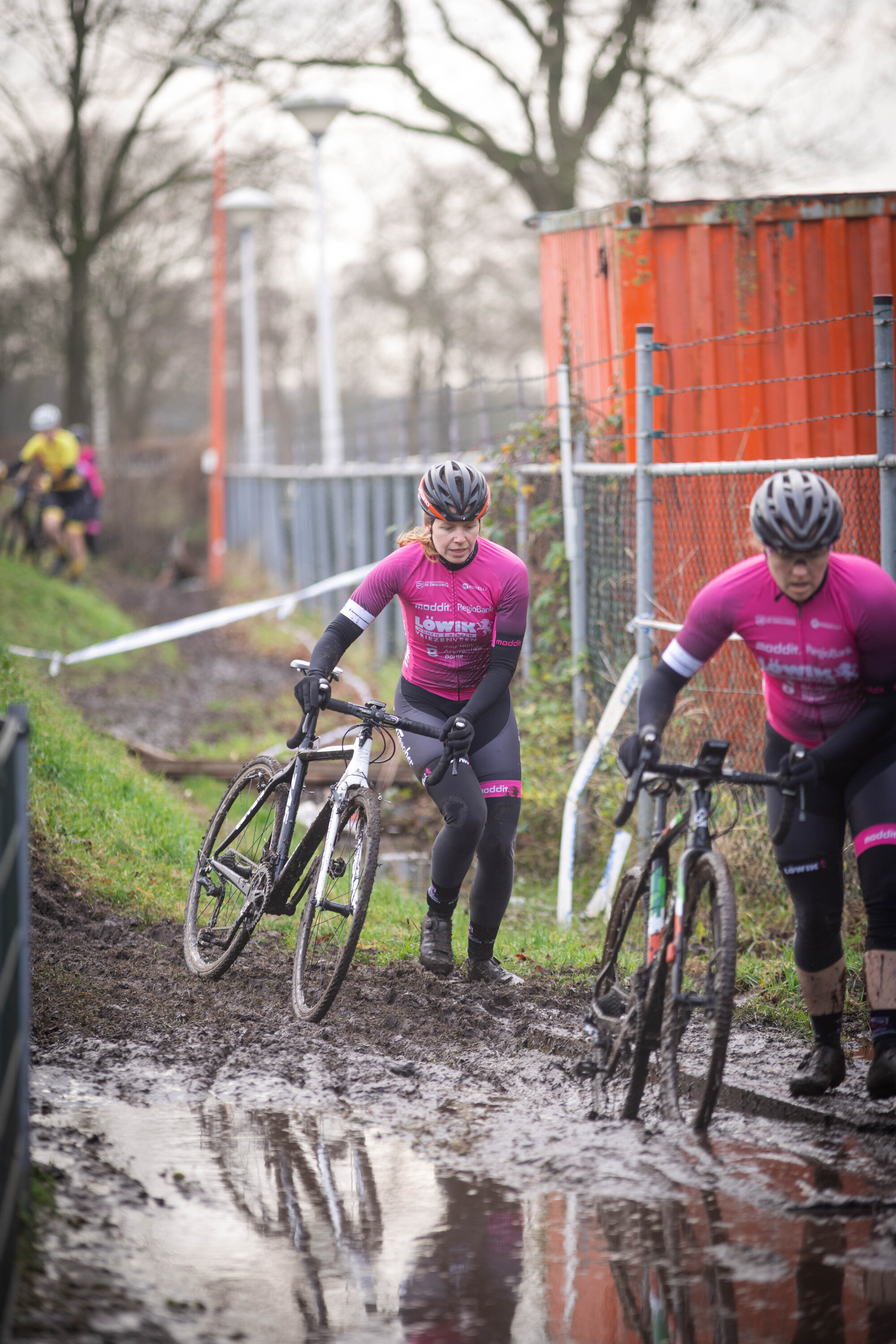 Two cyclists in pink and black outfits race on a muddy track.