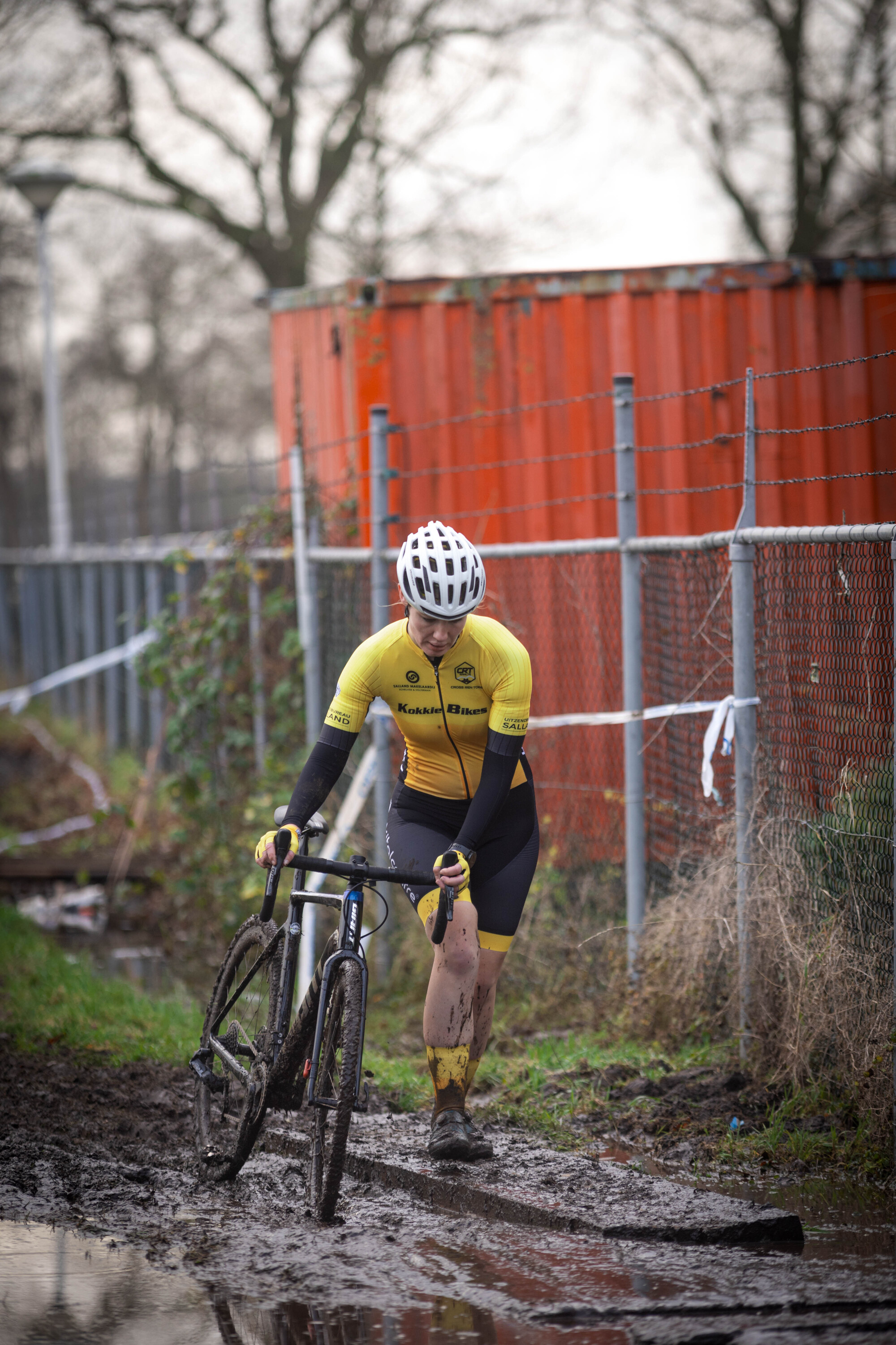 A cyclist wearing a yellow shirt and black shorts rides a bike on muddy ground.