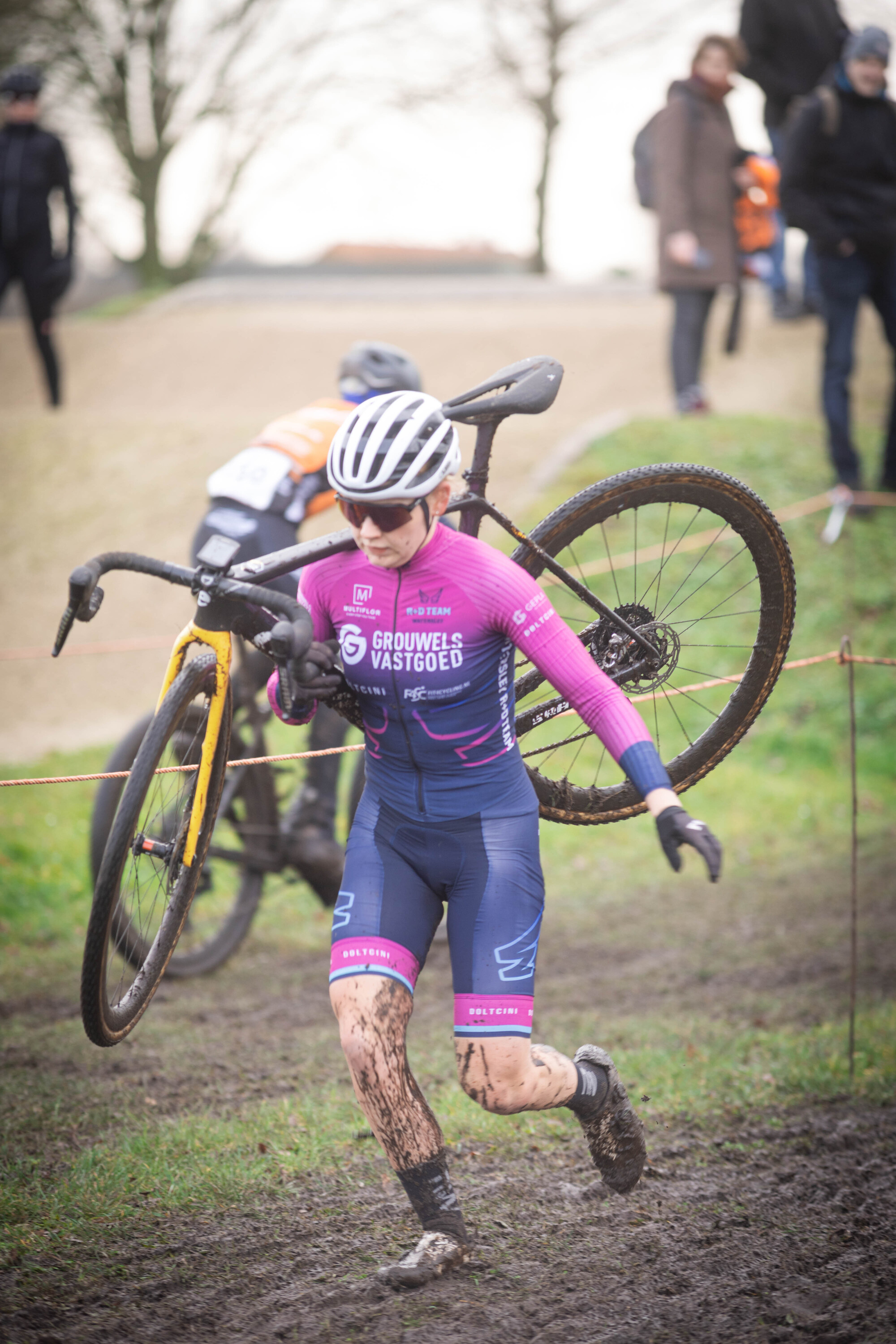 A young person in a blue and pink cycling uniform carries their bike over their shoulder on a muddy track.