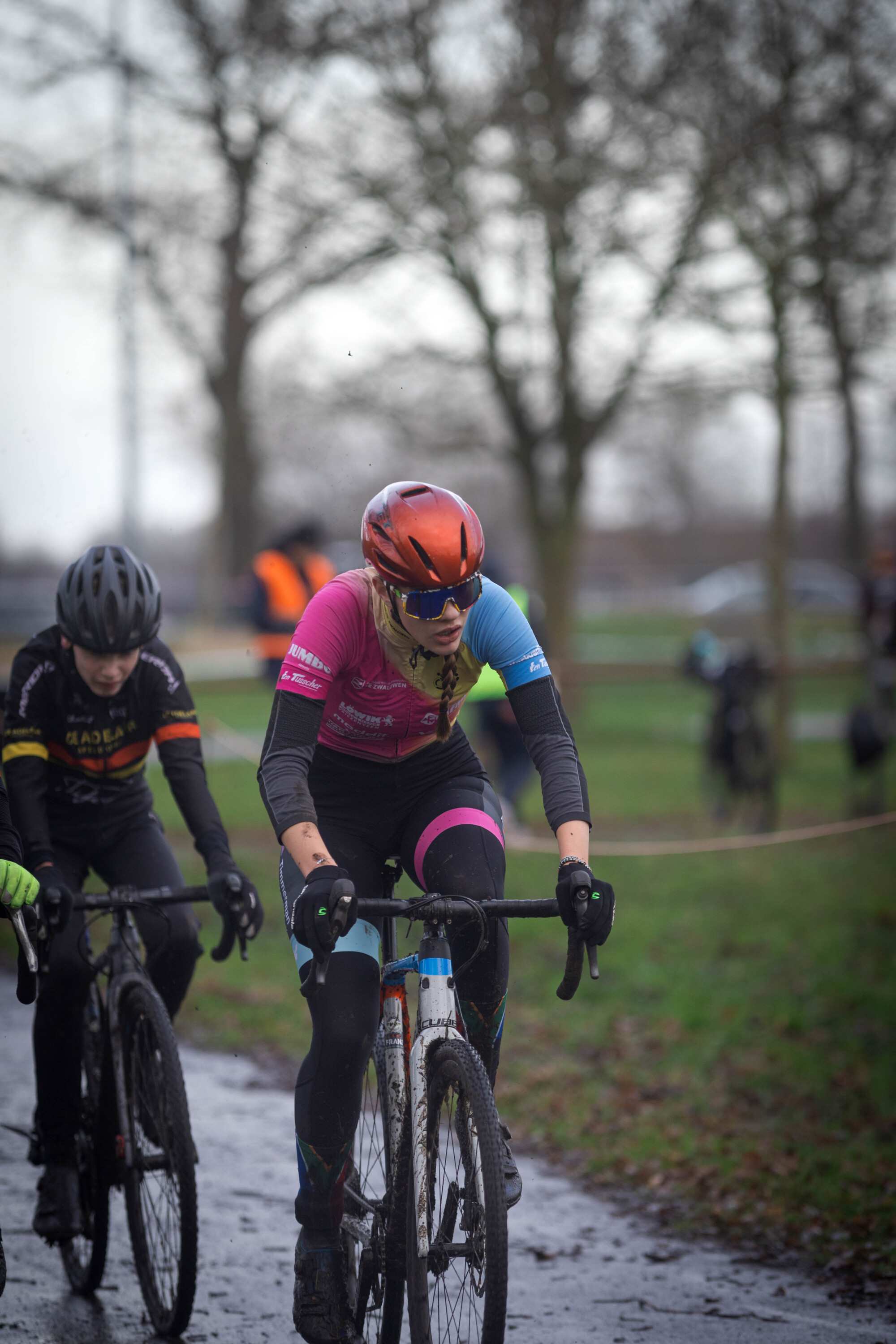 Two people are on bicycles, one wearing a pink shirt and the other an orange helmet.