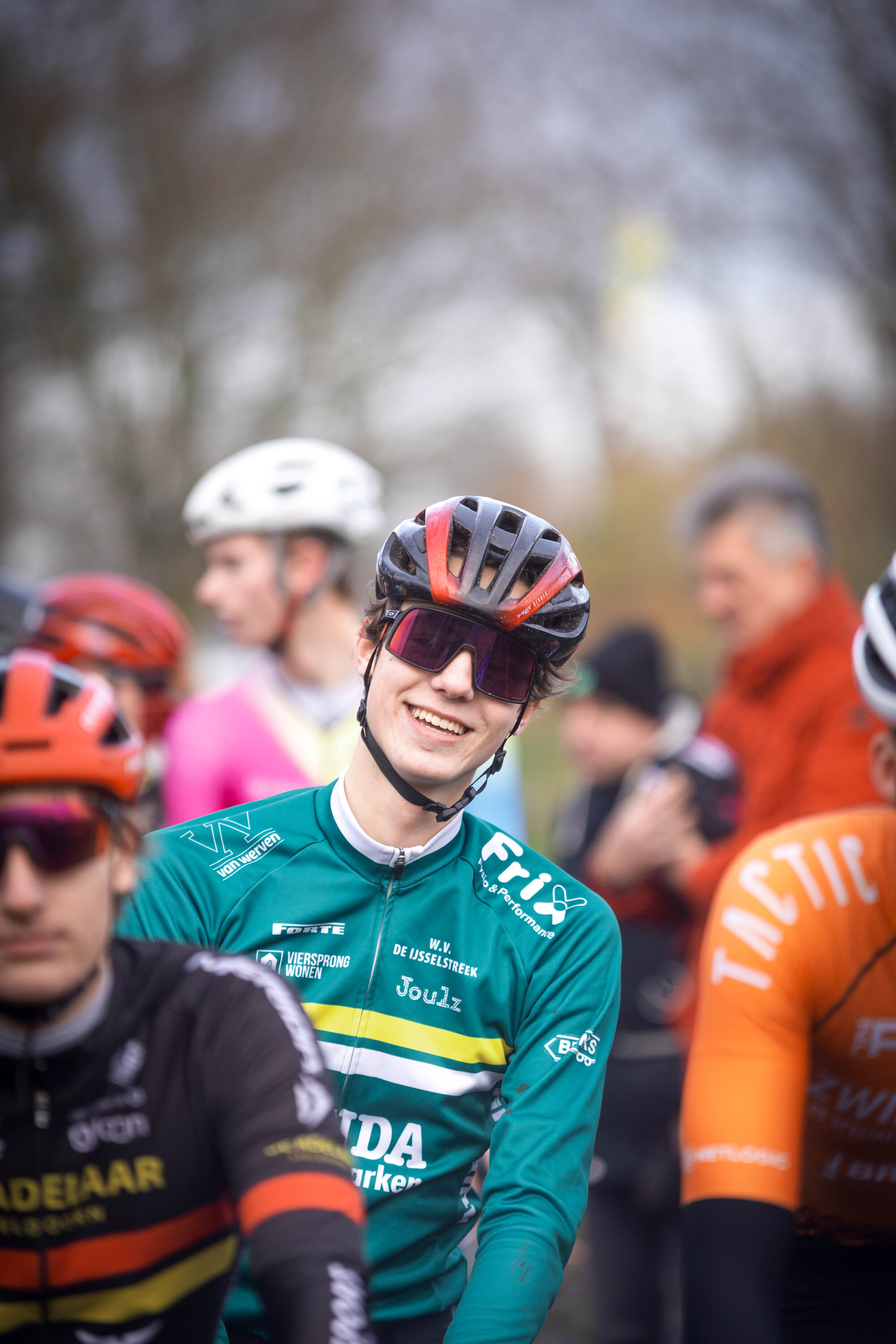 A group of cyclists wearing different colored uniforms are smiling and looking towards the camera.