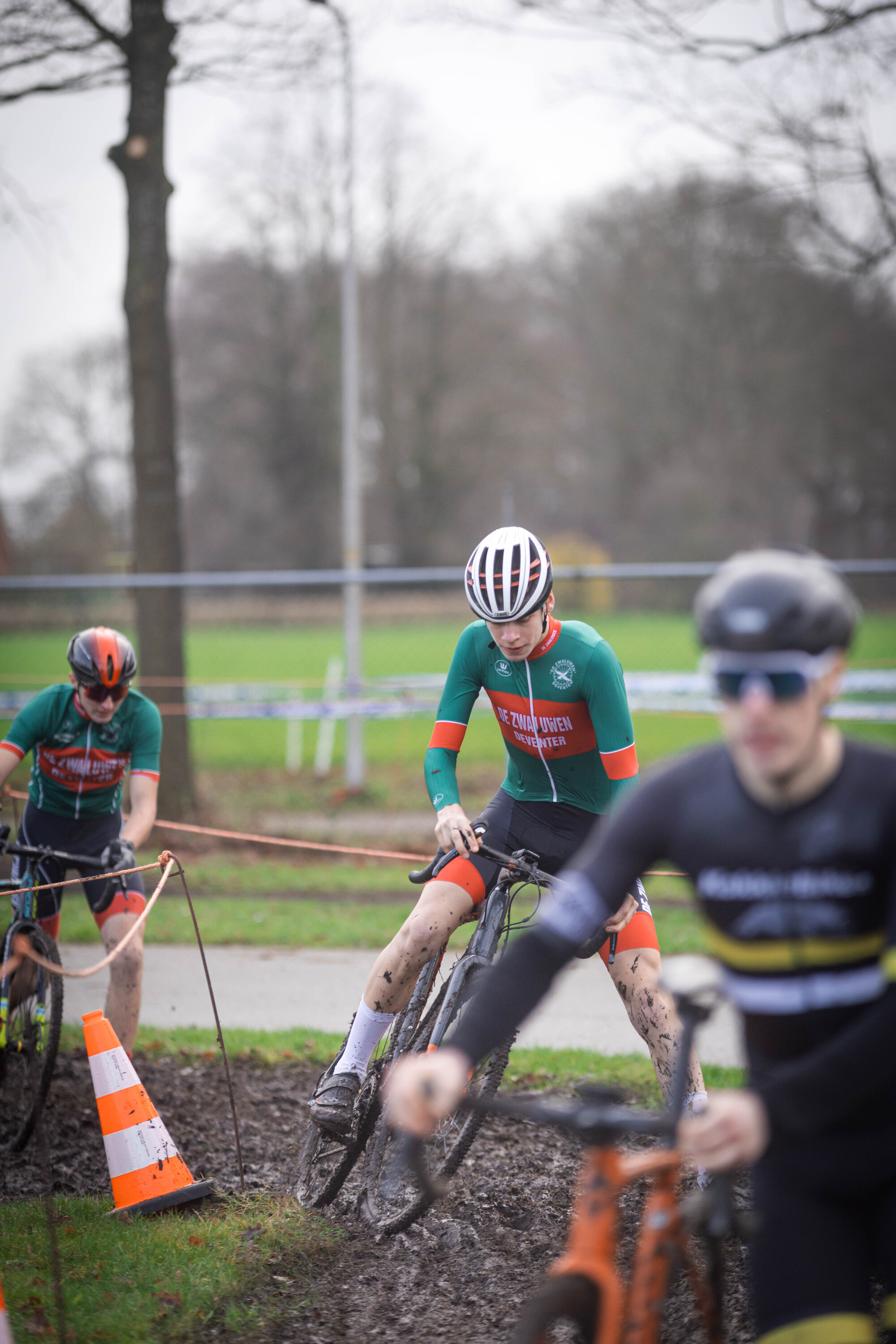 Three men are in a cyclocross race. The man in the green jersey is leading and wearing number 44.