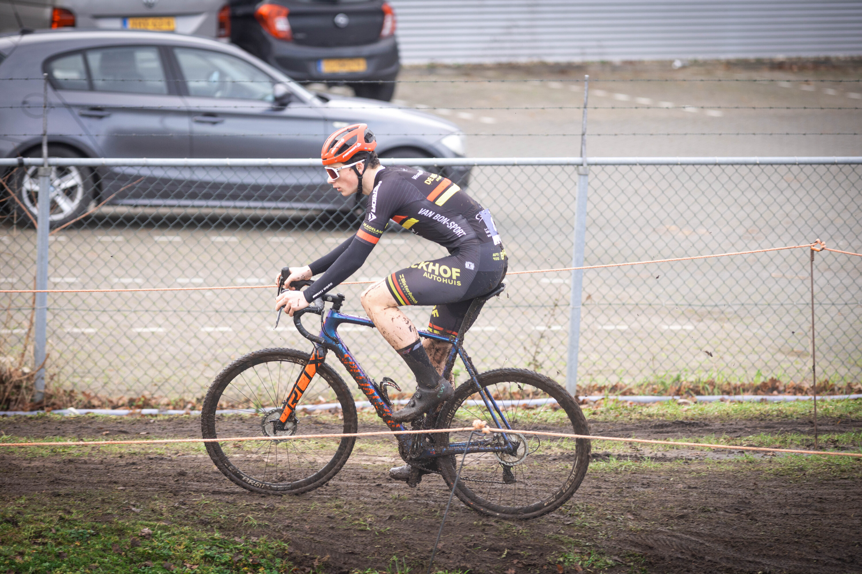 A man riding a bicycle on dirt, wearing Ju/El/B/E/A/M and an orange helmet.