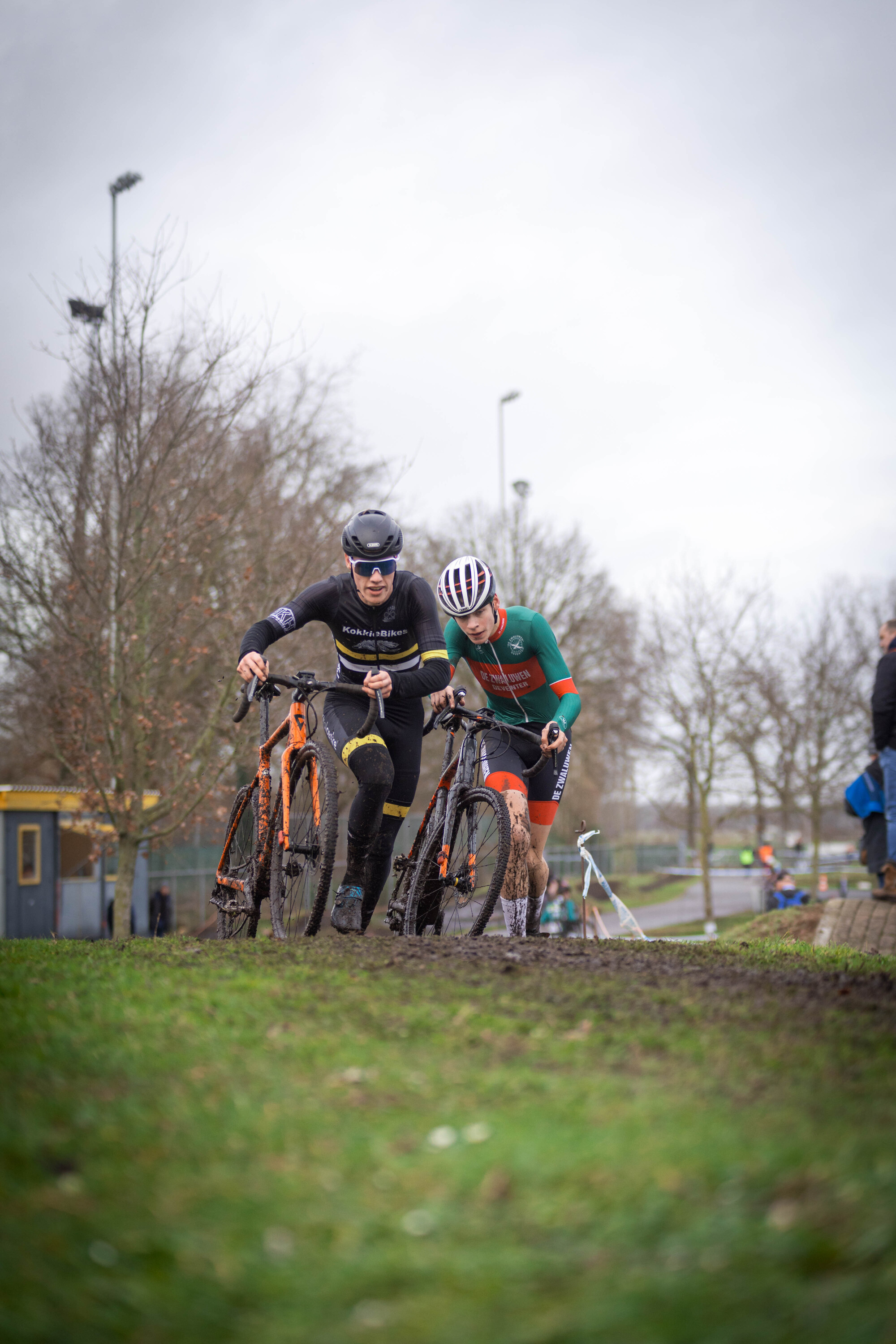 Two cyclists wearing black helmets are riding through mud.