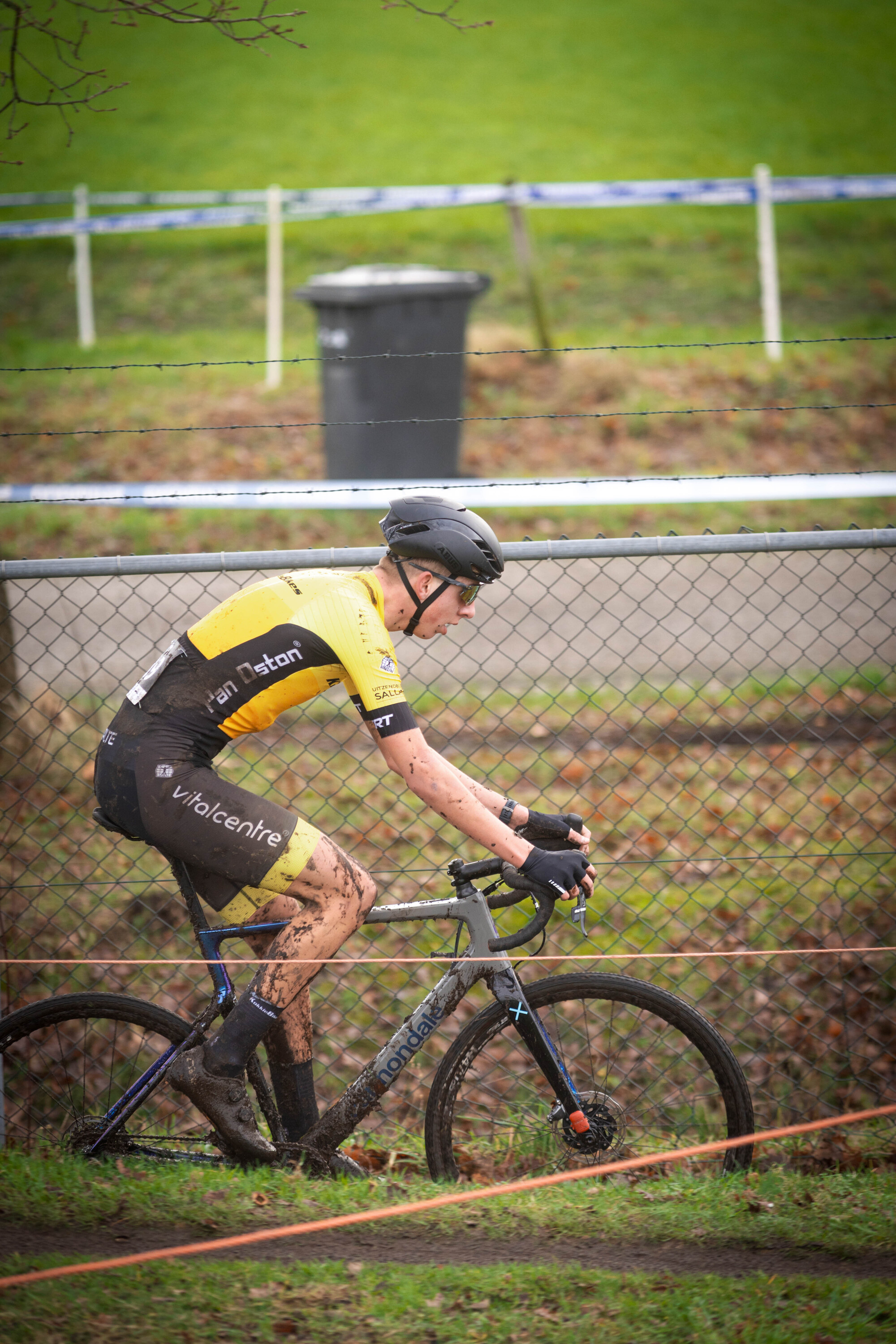 A cyclist in a yellow jersey with black lettering is riding his bike.