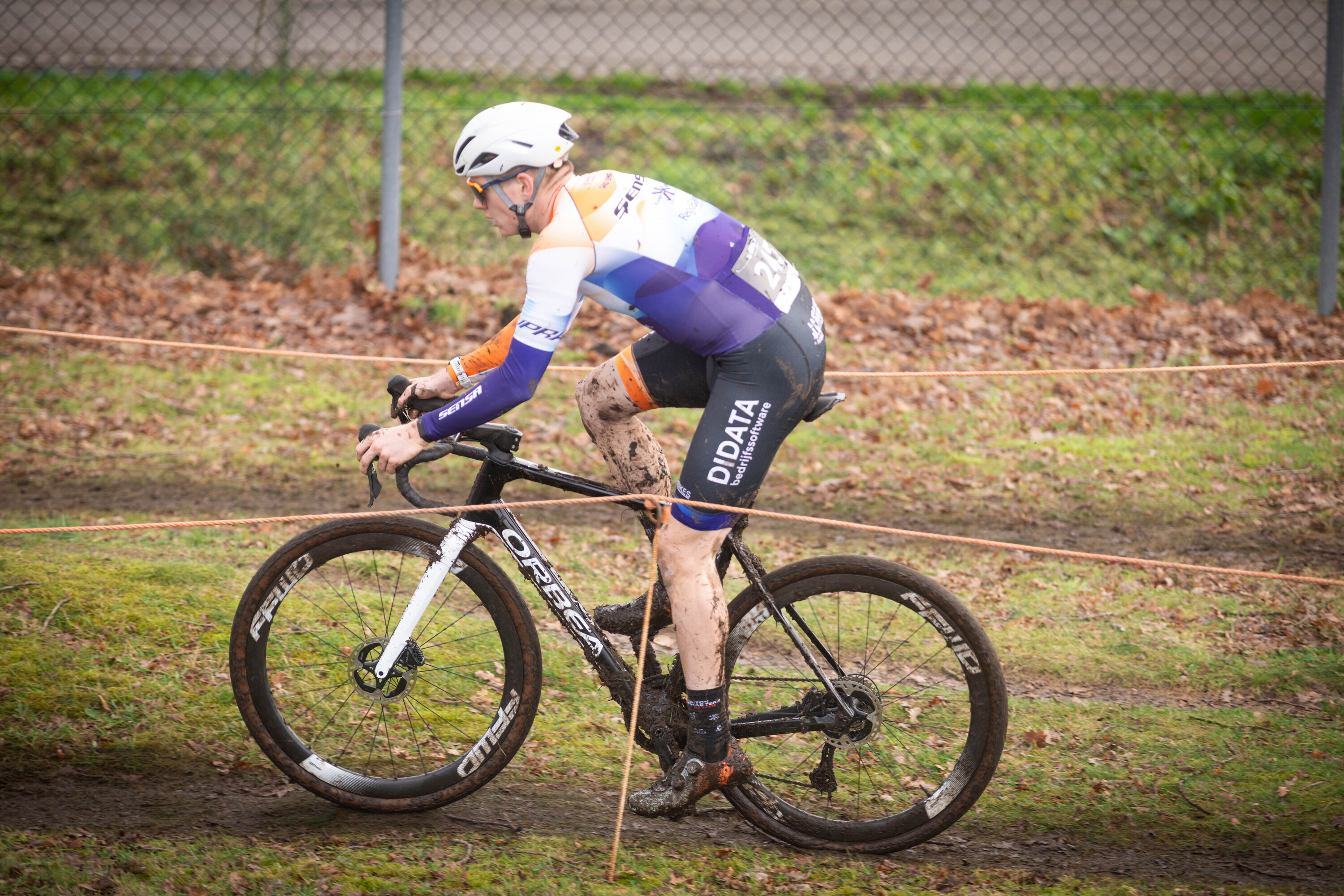 A man in a helmet and cycling gear is riding a black bike with orange wheels on the ground.