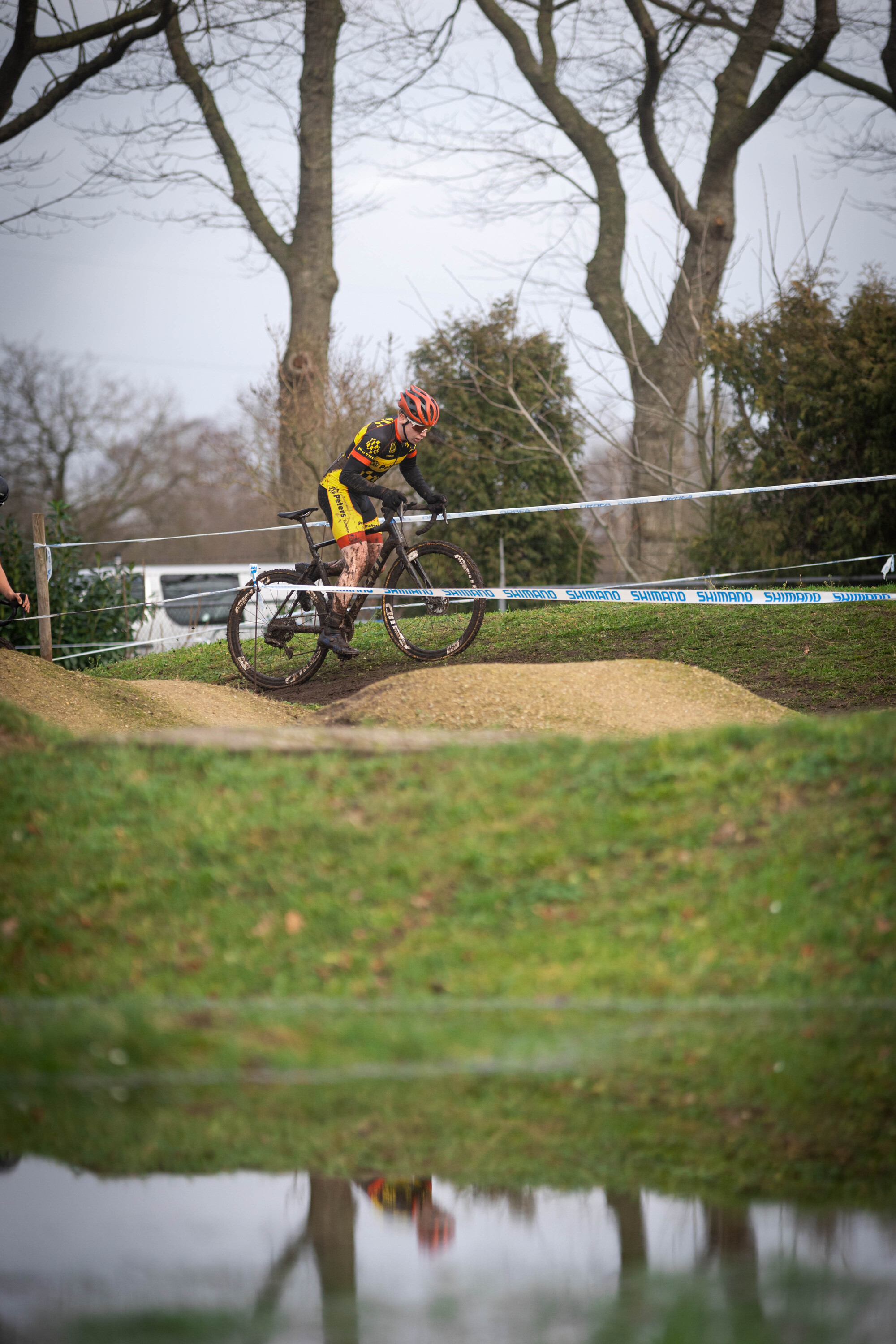 A cyclist, wearing a yellow and black shirt and an orange helmet, is going over a small jump on his bike.