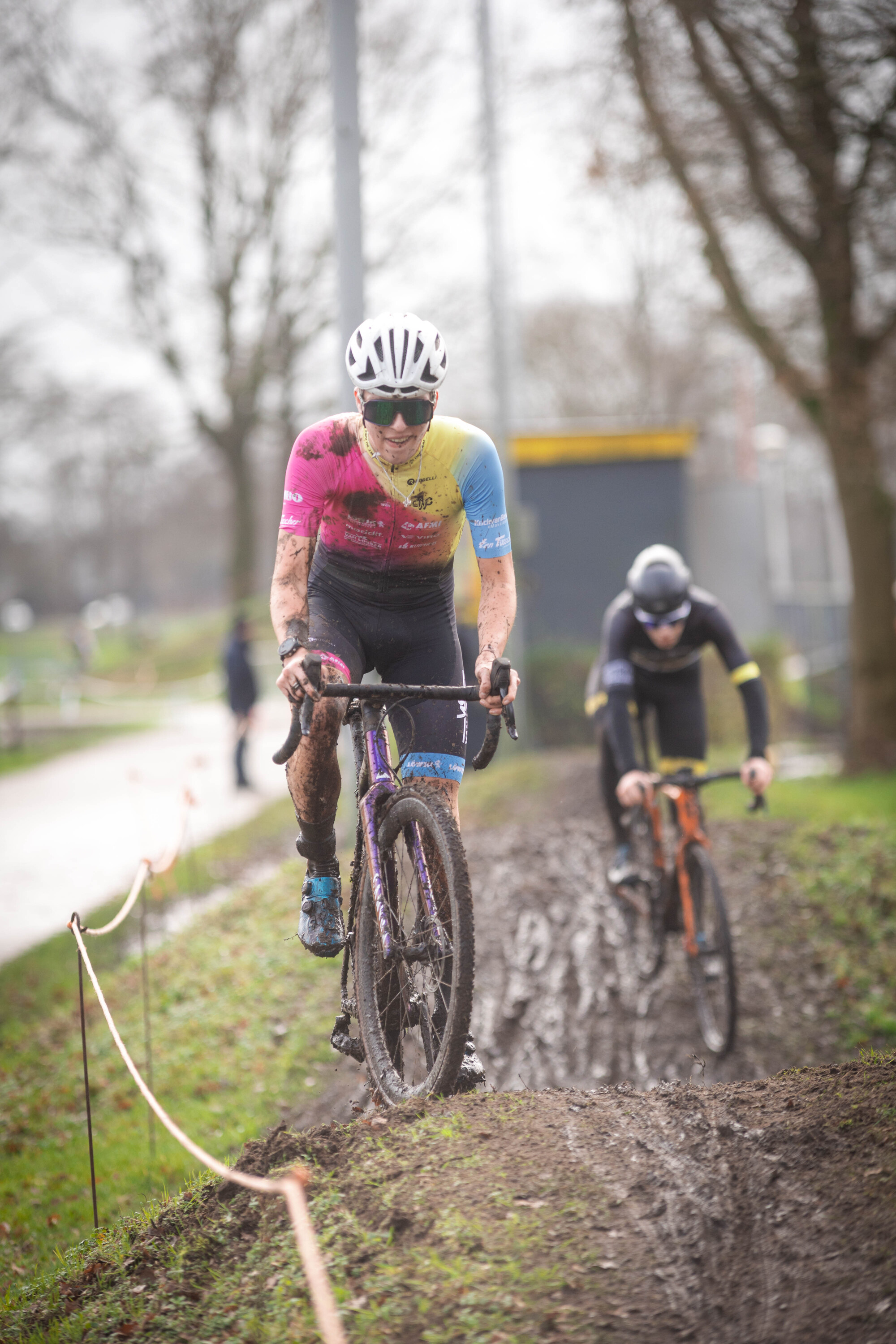 Two cyclists on muddy path with one wearing blue and yellow jersey.