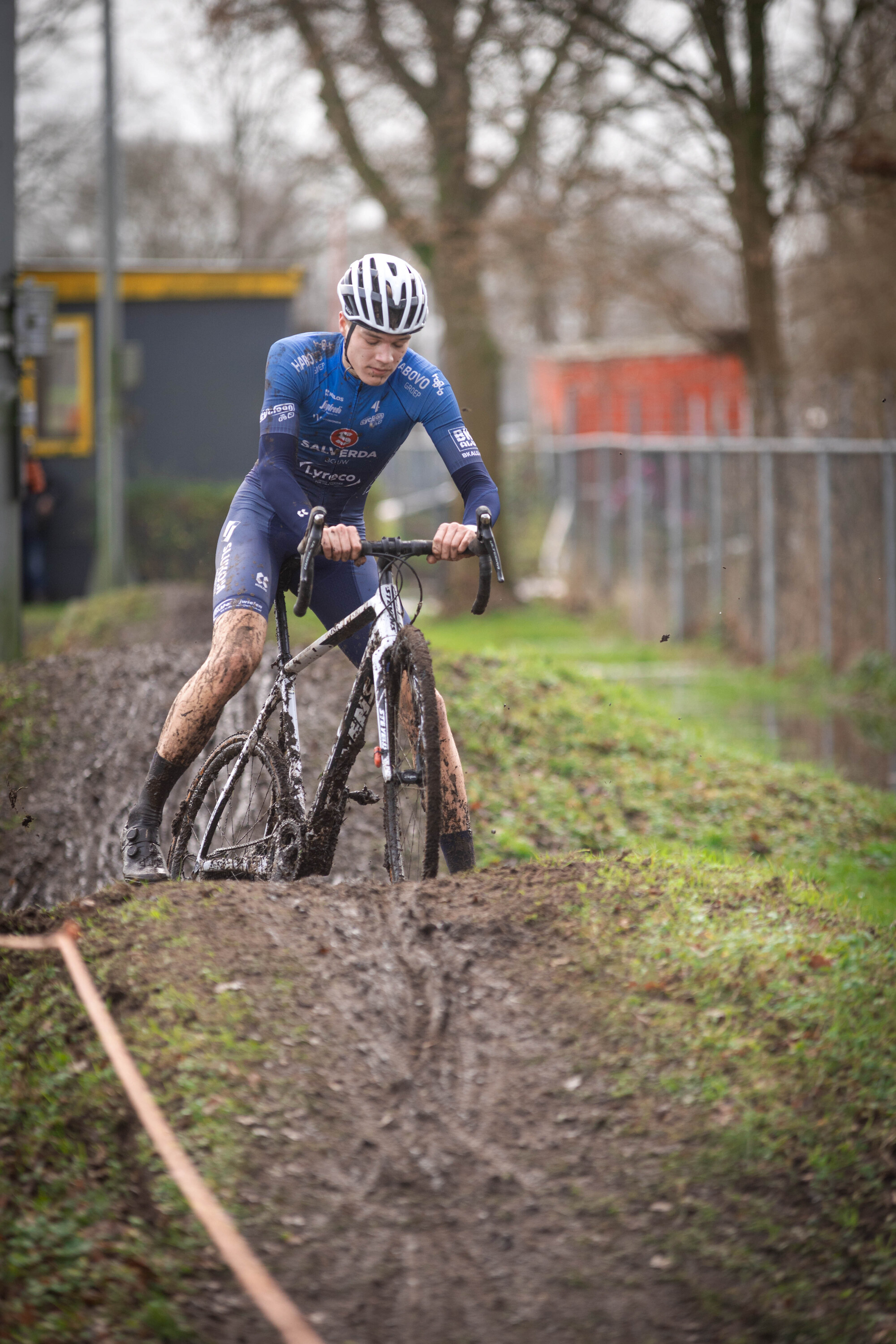 A person in a blue and white cyclocross uniform rides their bike through mud.