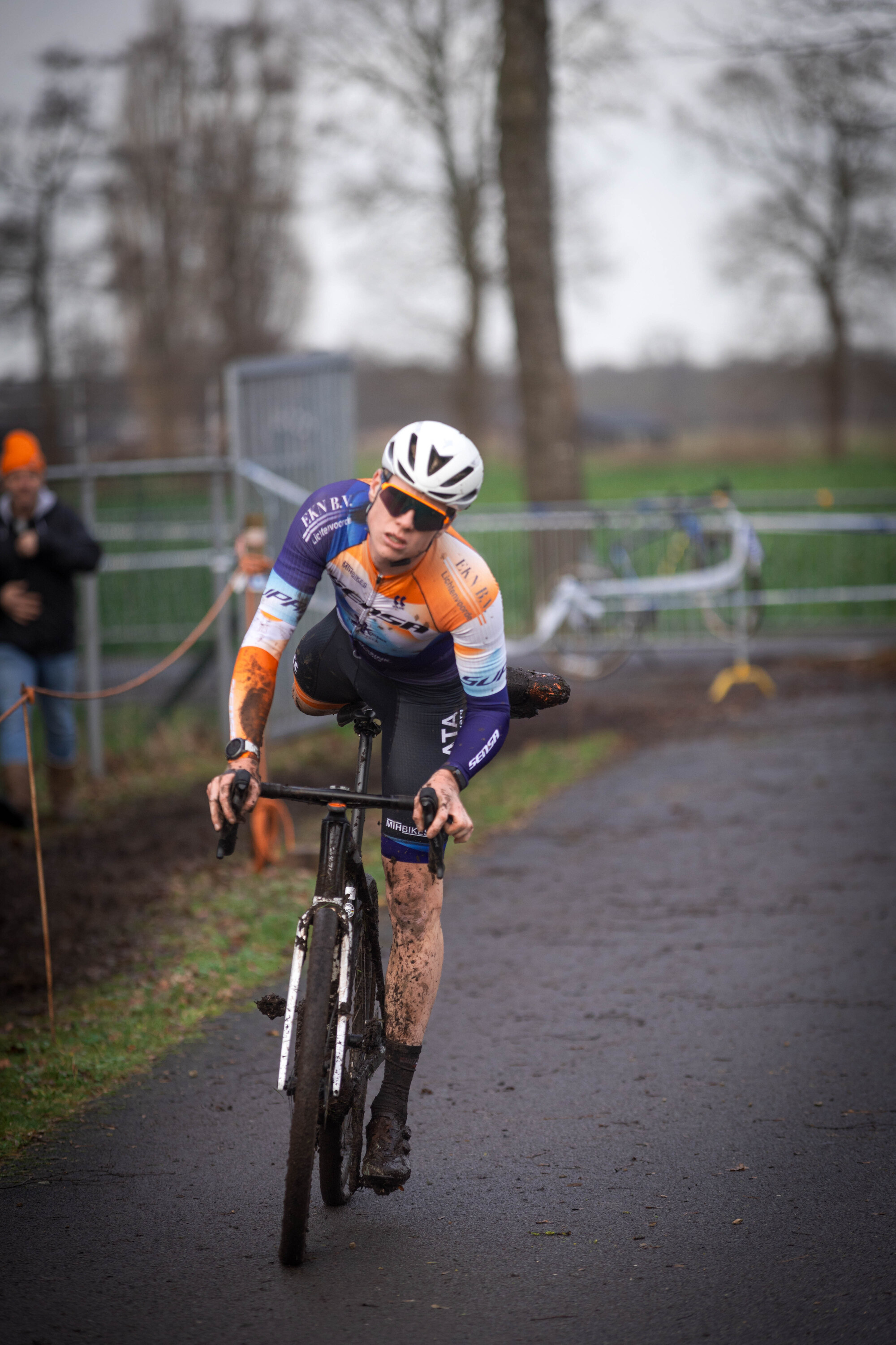 A man in a purple, orange, and white jersey riding his bike with mud on it.