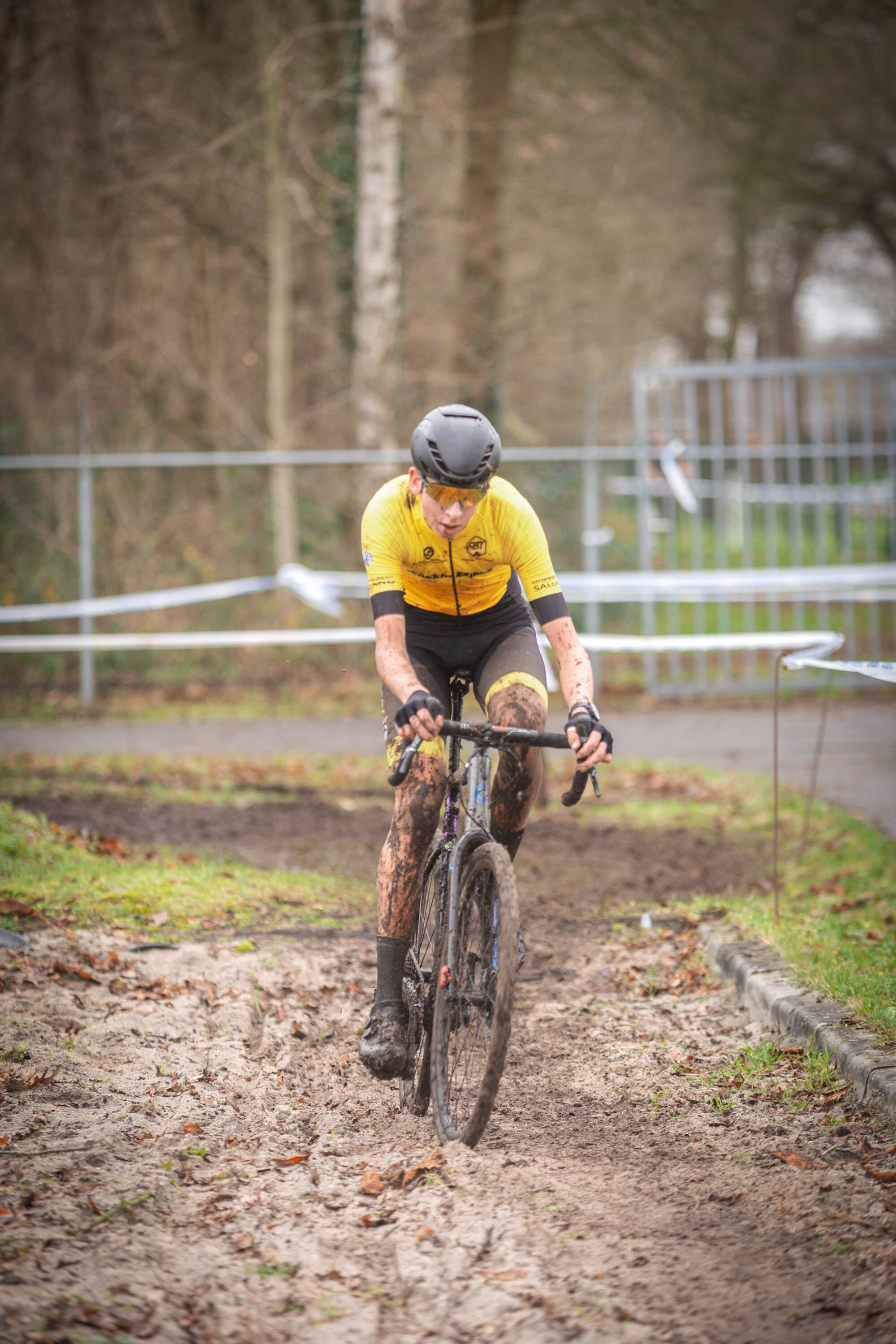 A cyclist is riding a bicycle on a path with trees in the background. The cyclist is wearing a yellow jersey and black pants.