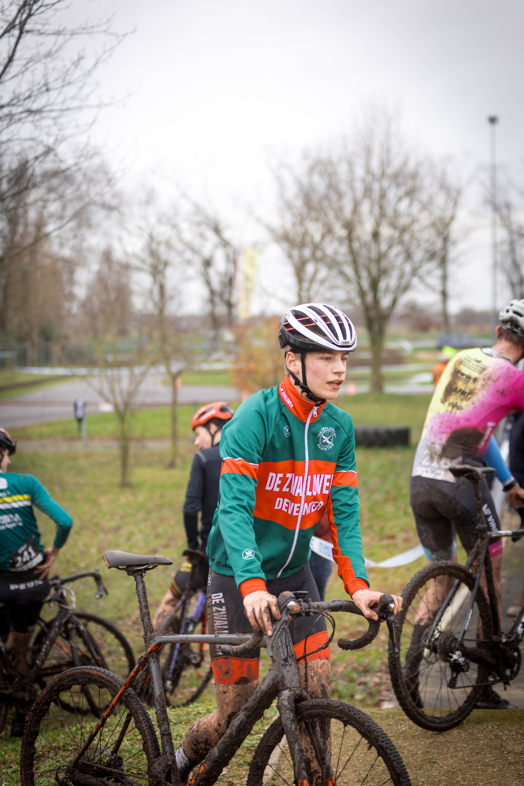 A group of cyclists in a grassy area, one wearing a green and orange jacket with the words "To live is to race".