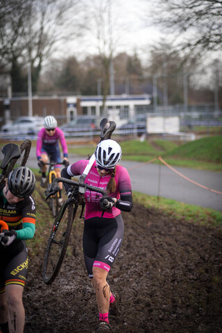 A woman in a red top and pink shorts is riding a bicycle through mud with two other people on bikes behind her.