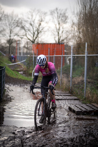 A bicyclist wearing a pink jersey and helmet riding through mud on a wet path.