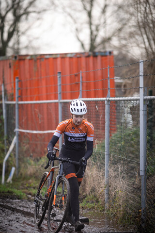 A woman wearing a orange and white jersey is riding a bicycle near some red containers.