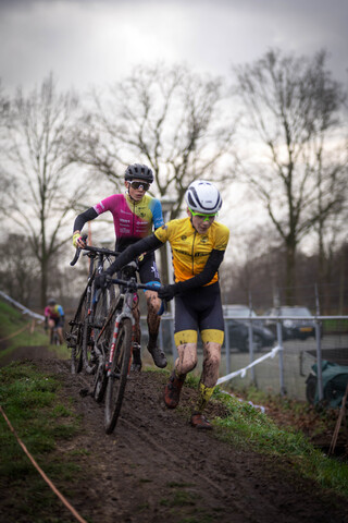 A man in a pink shirt is on a bike with two other men in yellow. They are crossing a muddy area.