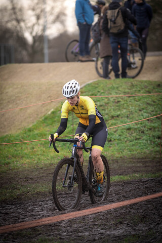 A cyclist in a yellow and black jersey is riding a dirty bike on a muddy track at a cyclocross race.