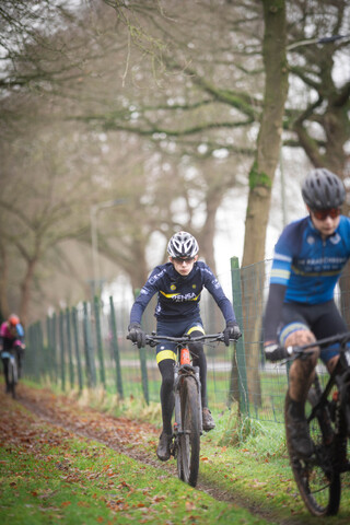 People ride bikes on a dirt path. The man in the blue shirt has the word "Grote" visible on his attire.