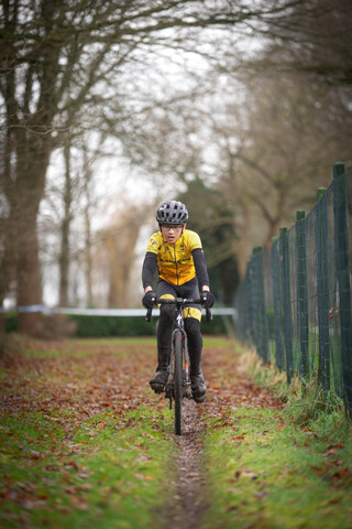 A young woman wearing a black and yellow top rides a bicycle down a path surrounded by trees.