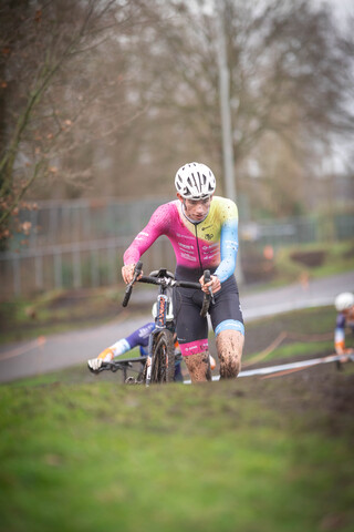 A man wearing a pink and blue jersey is riding his bike across the finish line at a cyclocross race.