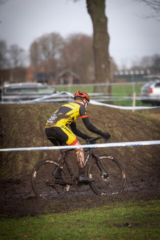 A man in a red and yellow suit is riding a black bicycle on muddy grass with the number 2023 on his back.