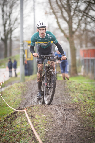 A man on a muddy trail on his bike with a green and yellow shirt that says Ju/El/Bc/Am.