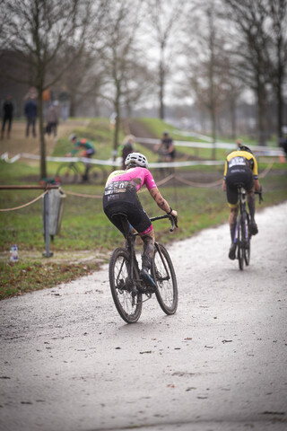 Two cyclists race on a road with trees in the background and one cyclist wearing a pink top.