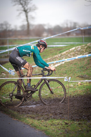 A man in a green and black outfit is riding a bicycle on muddy terrain, sponsored by Gow Raalte.