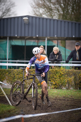 A man wearing a helmet and an orange, black, and white cycling outfit standing next to his black and white mountain bike.