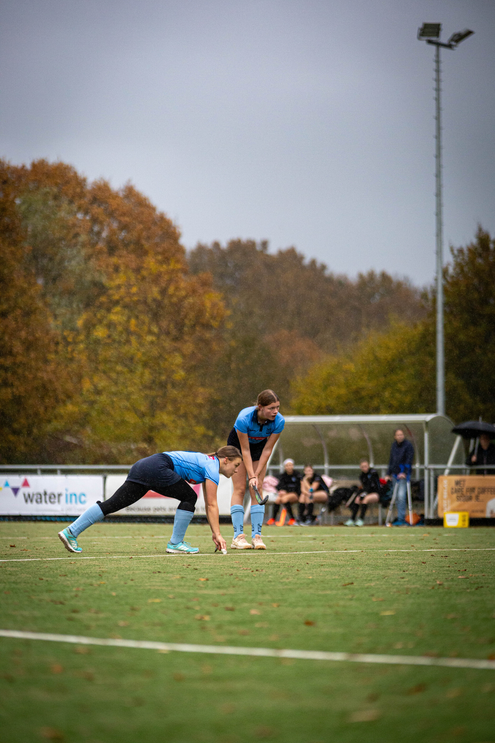 Two women in blue uniforms are playing hockey on a field.