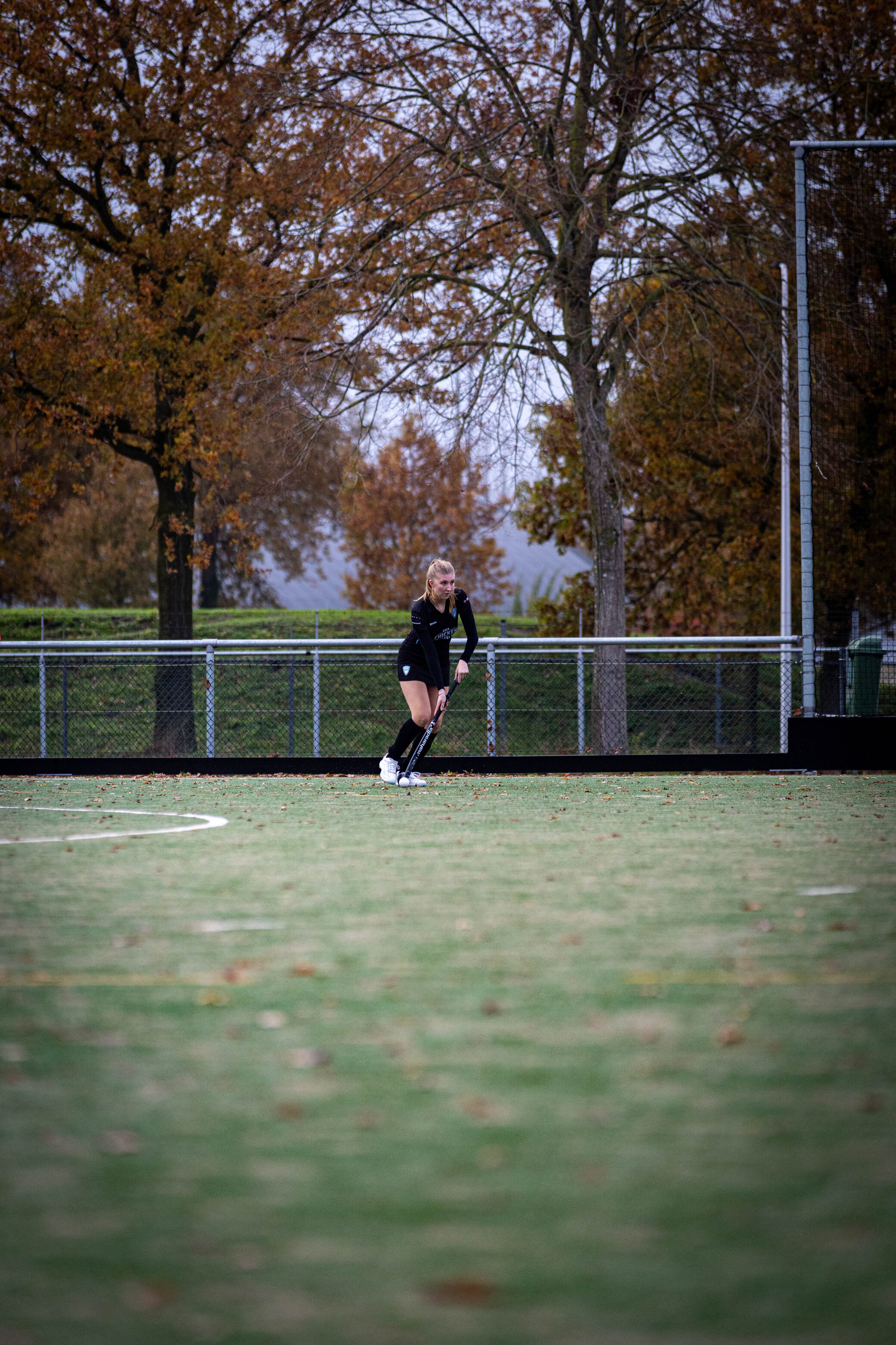 A woman in a black shirt and shorts is playing hockey on an empty field.