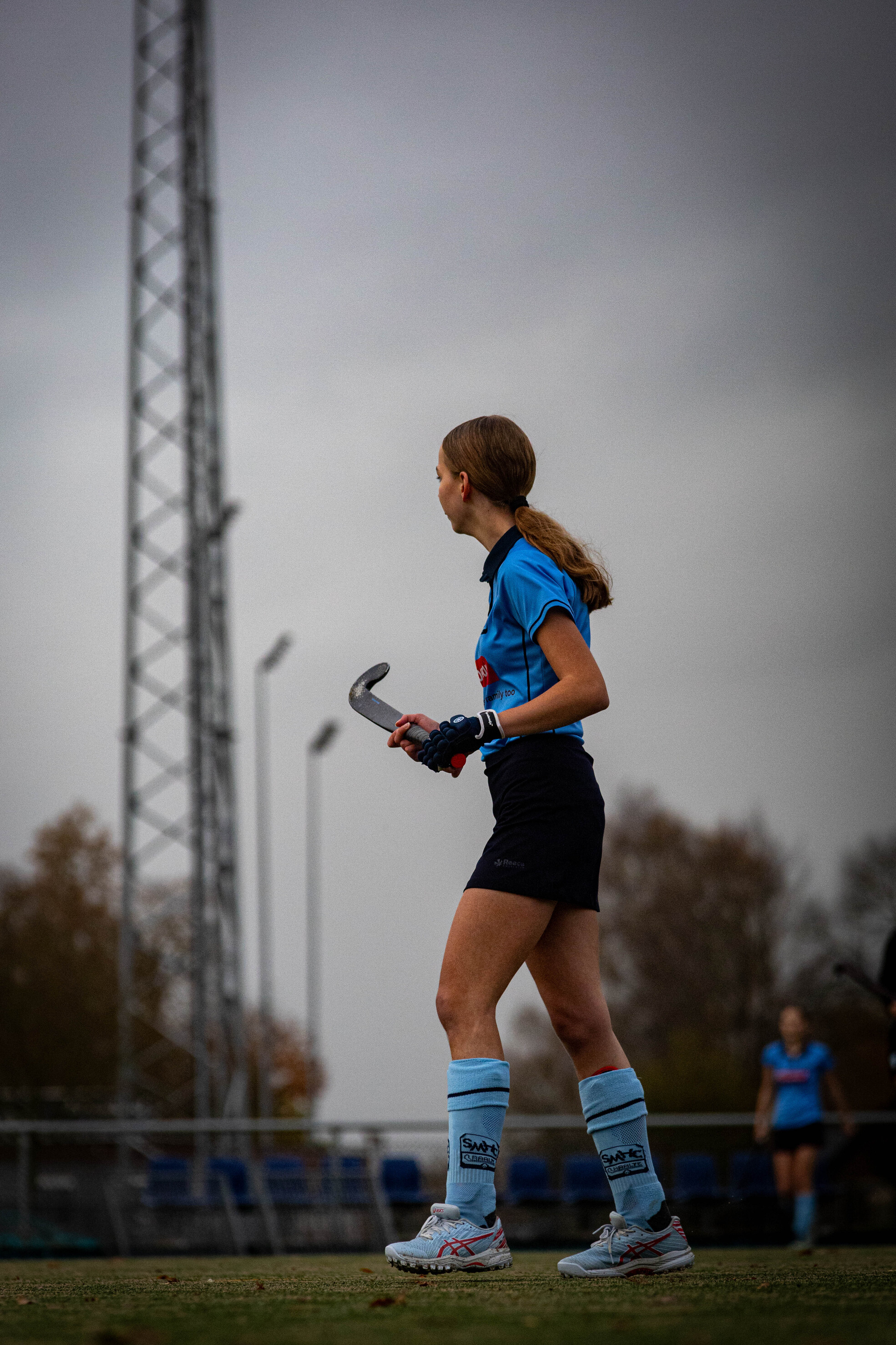 A female hockey player wearing blue and white uniform is walking on a field with her stick.