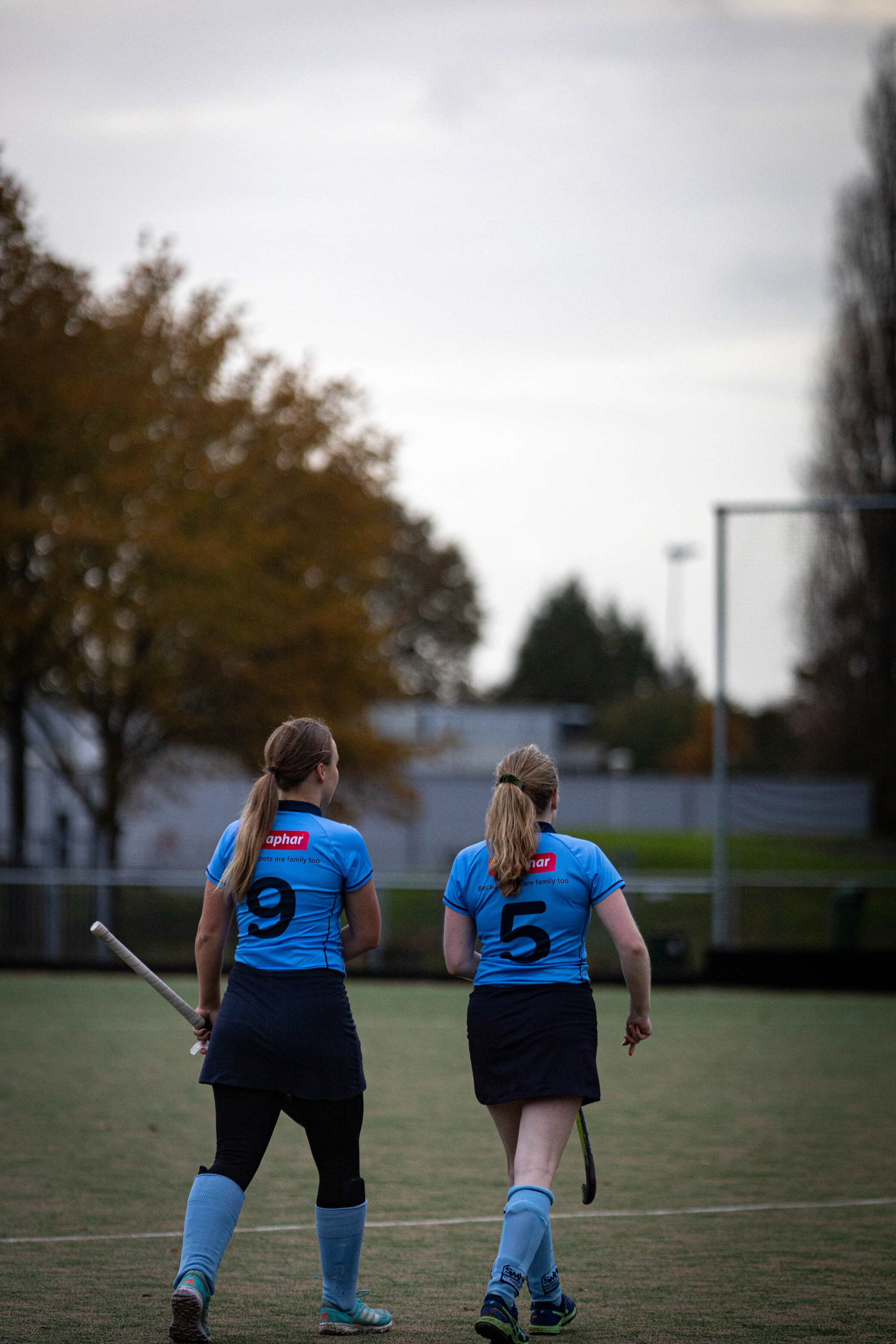 Two women in blue hockey jerseys stand on a field wearing SMHC 19-11.