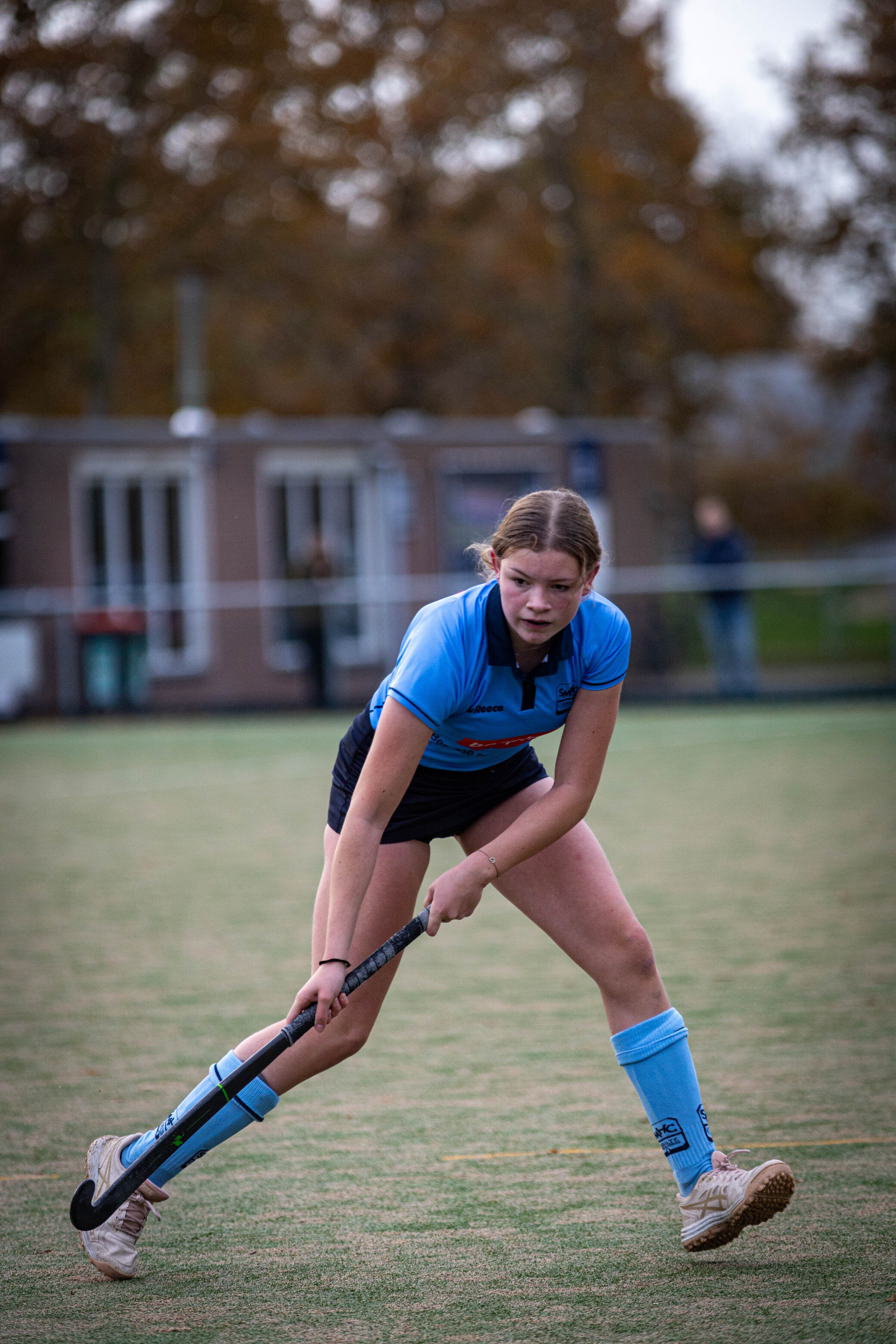 A young woman in a blue sports uniform on a field of grass, wearing the number 20, playing hockey.